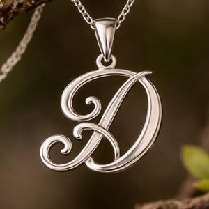 Peut inclure: Un collier pendentif argenté avec la lettre "D" en cursive. Le pendentif est suspendu à une fine chaîne. L'arrière-plan est une vue floue d'une branche d'arbre avec des feuilles vertes.
