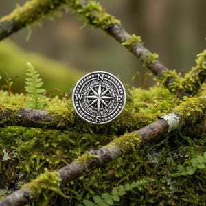 May include: A silver-colored ring with a compass design. The ring features a detailed compass rose with the cardinal directions (N, E, S, W) and a star in the center. The ring is set against a backdrop of moss-covered branches and greenery.