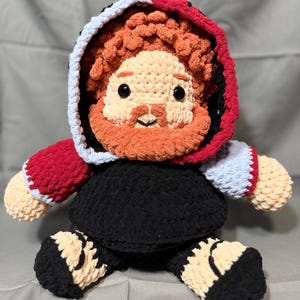 May include: A handmade crochet plush doll featuring a figure with curly orange hair and a matching beard. The doll wears a hooded garment with a black body, red and light blue sleeves, and a hood lined in red and light blue. It has black eyes.