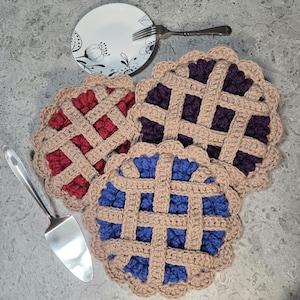 Op de afbeelding: Drie gehaakte onderzetters in de vorm van een taart, in rood, paars en blauw, elk met een bruin rooster. Een wit bord met bloemmotief en een vork zijn op de achtergrond.