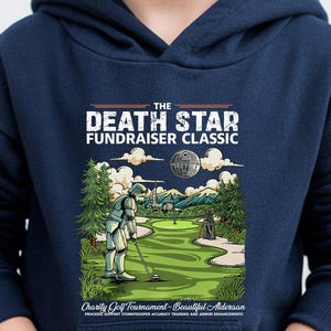 May include: Navy blue hooded sweatshirt featuring a graphic of a golf course with a Death Star theme. The design includes the text "THE DEATH STAR FUNDRAISER CLASSIC" and a golf-themed illustration. The sweatshirt has a front pocket.