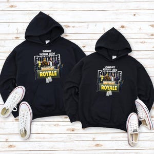 Pu&ograve; includere: Due felpe con cappuccio nere con il logo Fortnite e le scritte "Daddy Victory Crew" e "Mommy Victory Crew". Le scarpe da ginnastica bianche con strisce rosse sono posizionate vicino alle felpe. Le felpe hanno una grafica "Birthday Royale".