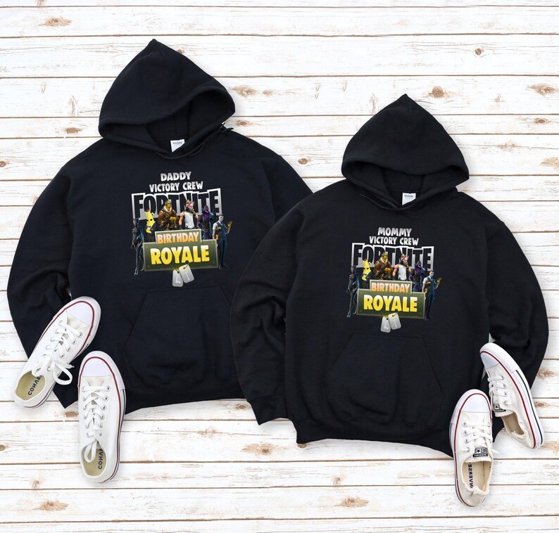 Pu&ograve; includere: Due felpe con cappuccio nere con il logo Fortnite e le scritte "Daddy Victory Crew" e "Mommy Victory Crew". Le scarpe da ginnastica bianche con strisce rosse sono posizionate vicino alle felpe. Le felpe hanno una grafica "Birthday Royale".