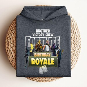 Pu&ograve; includere: Una felpa con cappuccio grigio scuro con la scritta "BROTHER VICTORY CREW FORTNITE BIRTHDAY ROYALE". Il design include personaggi dei cartoni animati. La felpa &egrave; esposta su una superficie circolare intrecciata.
