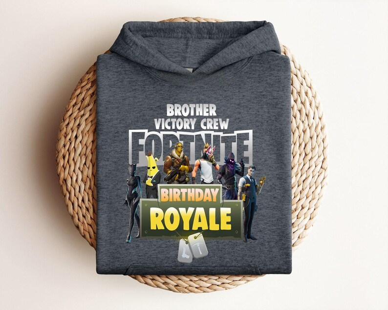Pu&ograve; includere: Una felpa con cappuccio grigio scuro con la scritta "BROTHER VICTORY CREW FORTNITE BIRTHDAY ROYALE". Il design include personaggi dei cartoni animati. La felpa &egrave; esposta su una superficie circolare intrecciata.