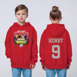 Pu&ograve; includere: Una felpa con cappuccio rossa di compleanno di Fortnite. La parte anteriore presenta il testo "BATTLE READY! FORTNITE BIRTHDAY ROYALE". Il retro mostra il nome "HENRY" e il numero "9" in grigio.
