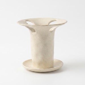 May include: A cream-coloured ceramic vessel with a speckled finish, featuring a cylindrical body, a wide circular base, and a flared top rim with three distinct cut-out openings that create a unique, open-structured silhouette for this object.