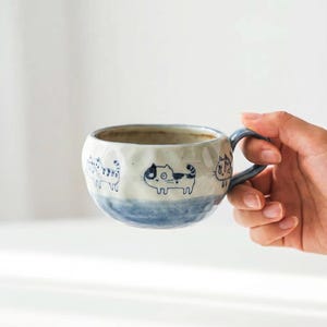 May include: A ceramic mug with a textured, dimpled surface featuring hand-painted blue cat illustrations. The mug has a rounded shape, a blue-glazed base, and a matching blue handle. The interior is off-white, and the exterior displays multiple cat figures.