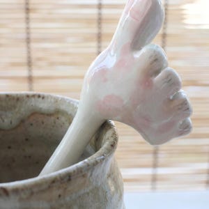 May include: A ceramic spoon shaped like a thumbs-up, with a white base and pink detailing. It sits in a textured, beige ceramic bowl. The background features a bamboo blind.