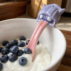 May include: A ceramic spoon with a purple monster head and a pink tongue, resting in a white bowl filled with yogurt and blueberries. The spoon's design is whimsical and unique, adding a playful touch to the breakfast setting.