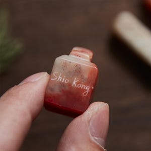 Puede incluir: Pequeña talla de piedra rectangular roja y blanca con forma de botella. El texto "Shio Kong" está grabado en la parte delantera. La talla se sujeta entre dos dedos.