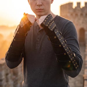 May include: A pair of black forearm bracers featuring multiple horizontal leather straps and metallic studs. The bracers extend from the wrist to the mid-forearm, secured with adjustable buckles and rivets, worn over a dark gray long-sleeve shirt.