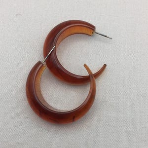 May include: A pair of brown hoop earrings. The earrings have a crescent shape with a lighter brown interior. The earrings are made of a translucent material and have a silver post.
