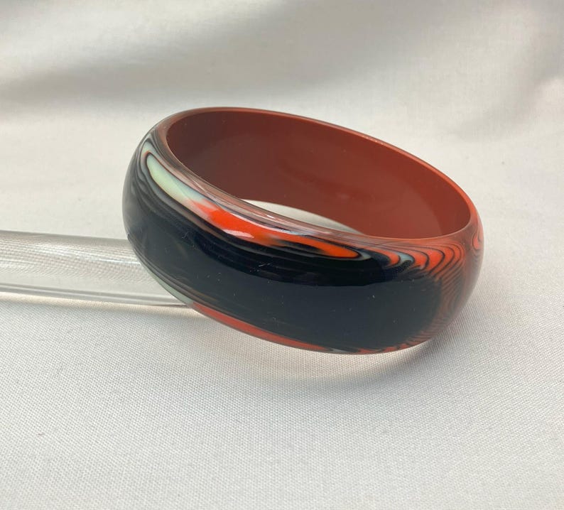 May include: A wide, round bangle bracelet with a marbled design. The bracelet features a dark red interior and a black, orange, and white exterior. The bracelet is made of a glossy material and is displayed on a clear stand.
