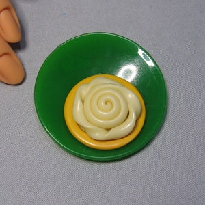 Peut inclure: Une rose en plastique blanche avec un centre jaune, posée sur une soucoupe verte et jaune.