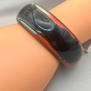May include: A wide, marbled bangle bracelet in shades of black, orange, and white. The glossy surface reflects light, highlighting the swirling patterns. The bracelet is a bold accessory.