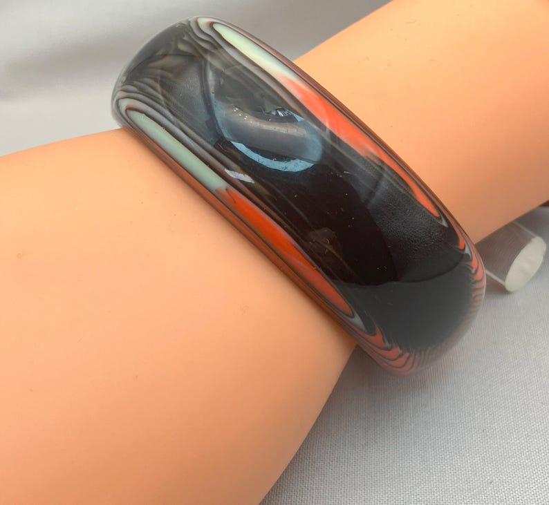 May include: A wide, marbled bangle bracelet in shades of black, orange, and white. The glossy surface reflects light, highlighting the swirling patterns. The bracelet is a bold accessory.