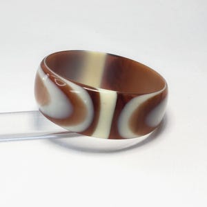 May include: A wide, marbled bangle bracelet in shades of brown and cream. The bracelet has a rounded shape with a smooth, glossy finish. The design features swirling patterns of light and dark colors, creating a unique accessory.