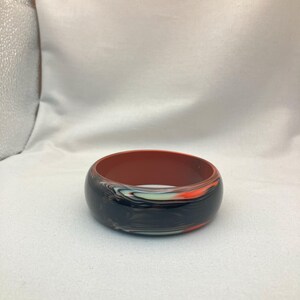 May include: A wide, round bangle bracelet with a marbled design. The exterior features a mix of black, orange, and white swirls, while the interior is a solid reddish-brown color. The bracelet is a bold accessory.