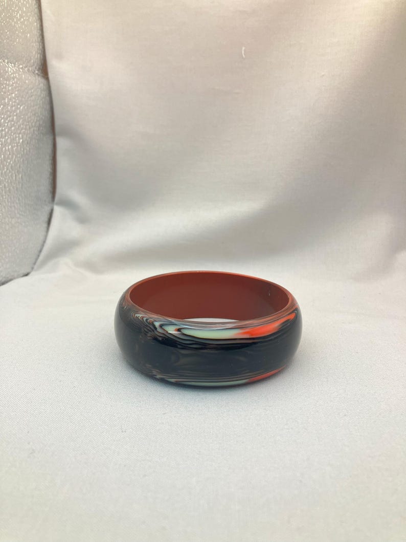 May include: A wide, round bangle bracelet with a marbled design. The exterior features a mix of black, orange, and white swirls, while the interior is a solid reddish-brown color. The bracelet is a bold accessory.