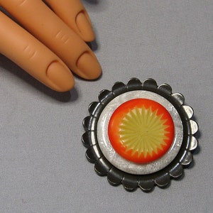 Peut inclure: Broche de style vintage avec un design en couches. Elle présente un motif central en forme d'éclat de soleil orange et jaune, posé sur un fond blanc texturé et encadré par une bordure en métal foncé festonnée. Un accessoire unique.