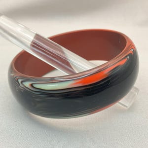 May include: A wide, marbled bangle bracelet in shades of black, orange, white, and pale green. The interior is a solid reddish-brown. A clear, cylindrical object rests inside the bracelet.
