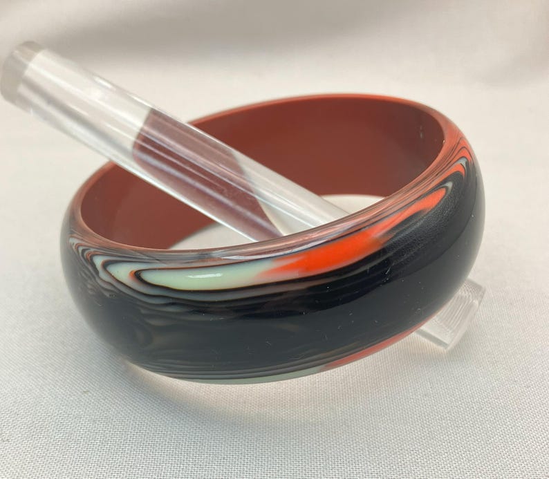 May include: A wide, marbled bangle bracelet in shades of black, orange, white, and pale green. The interior is a solid reddish-brown. A clear, cylindrical object rests inside the bracelet.