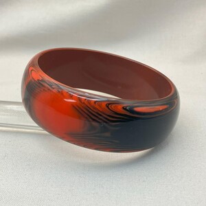 May include: A marbled bangle bracelet in shades of red, black, and brown. The wide, rounded bracelet has a glossy finish and a swirling pattern. The interior of the bracelet is a solid brown color. A stylish accessory.