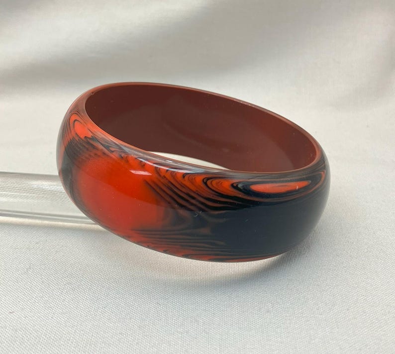 May include: A marbled bangle bracelet in shades of red, black, and brown. The wide, rounded bracelet has a glossy finish and a swirling pattern. The interior of the bracelet is a solid brown color. A stylish accessory.
