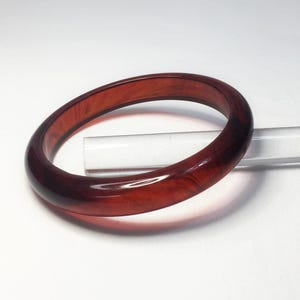 May include: A round, reddish-brown bangle bracelet. The bracelet has a smooth, polished surface and a marbled appearance. The jewelry is made of a translucent material, and the light reflects off the surface. The bracelet is a classic accessory.