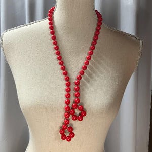 May include: A long, red beaded necklace displayed on a mannequin. The necklace features round, glossy beads and a unique, looped design at the bottom. The beads are a vibrant, solid red color, creating a bold statement piece.