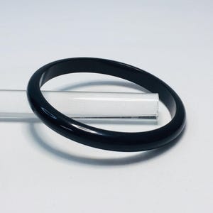 May include: A glossy, black bangle bracelet. The circular bracelet is made of a solid, dark material. The bracelet is resting on a clear, rectangular object against a white background. The bangle is a simple, elegant accessory.