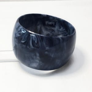 May include: A wide bangle bracelet featuring a polished finish with a marbled pattern in shades of dark charcoal gray, black, and white. The circular accessory has a smooth, rounded exterior surface and a hollow center, showcasing a swirling design.