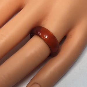 Op de afbeelding: Een roodbruine ring met een glad, gepolijst oppervlak. De ring is een effen band en wordt om een vinger gedragen. De kleur van de ring is een warme, aardse tint en lijkt van een doorschijnend materiaal te zijn gemaakt.