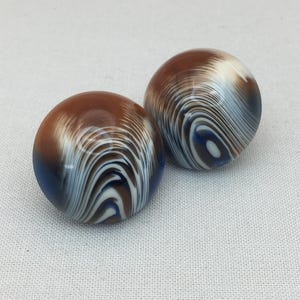 Può includere: Due perline rotonde in vetro marmorizzato. Le perline sono marroni, bianche e blu, con un motivo a vortice. Le perline sono su uno sfondo bianco.