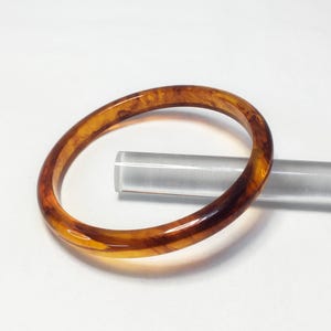 May include: A circular bangle bracelet featuring a polished, translucent amber-colored resin with swirling dark brown and golden-yellow marbled patterns throughout the material. The bracelet has a smooth, rounded surface and a glossy finish.