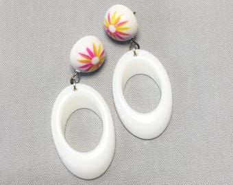 May include: A pair of drop earrings featuring a white, dome-shaped stud with a pink and yellow starburst pattern. Each stud is connected by a silver-toned metal link to a large, smooth, white oval-shaped hoop pendant with a hollow center.