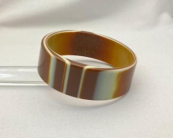 May include: A wide, striped bangle bracelet. The bracelet features alternating bands of brown, tan, and off-white. The bracelet is made of a smooth, glossy material. The bracelet is a fashion accessory.