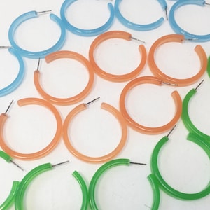 May include: A collection of circular hoop earrings in translucent blue, orange, and green colors. Each earring features a thin metal post for attachment. The hoops are arranged in a scattered pattern, showcasing their smooth, rounded plastic construction.