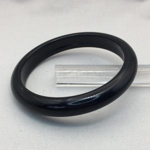 May include: A glossy, black bangle bracelet. The circular bracelet is made of a solid, dark material and has a smooth, polished surface. The bracelet is resting on a clear, rectangular support.
