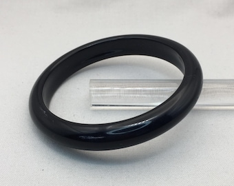 May include: A glossy, black bangle bracelet. The circular bracelet is made of a solid, dark material and has a smooth, polished surface. The bracelet is resting on a clear, rectangular support.