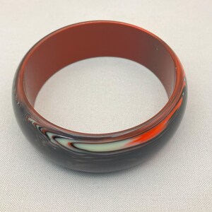 May include: A round bangle bracelet with a dark exterior and a reddish-brown interior. The exterior features a swirling pattern of black, white, and orange. The bracelet is made of a solid material.