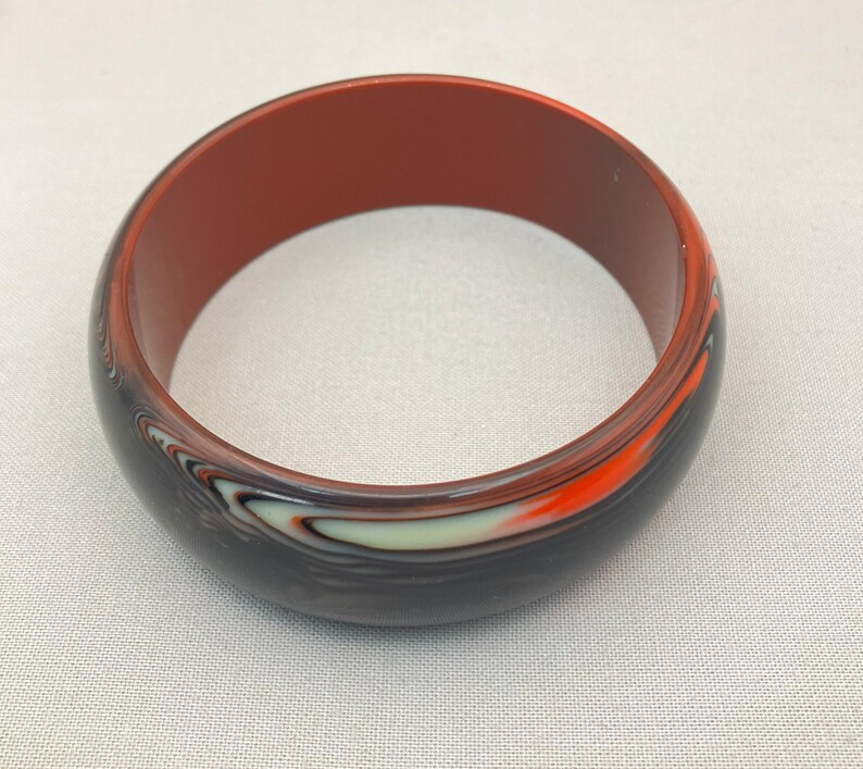 May include: A round bangle bracelet with a dark exterior and a reddish-brown interior. The exterior features a swirling pattern of black, white, and orange. The bracelet is made of a solid material.