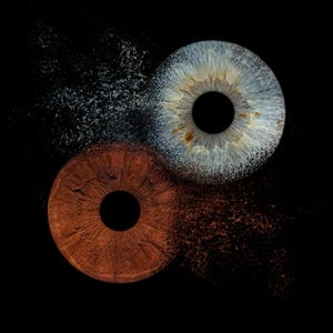 May include: A conceptual image of two human irises, one brown and one blue, set against a black background. The irises appear to be disintegrating into particles, creating a dynamic visual effect. The image evokes themes of duality and transformation.