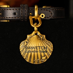 Puede incluir: Una placa dorada para mascotas con forma de concha, grabada con el nombre "PAWETCH" y un número de teléfono. La placa está sujeta a un collar de perro de cuero negro con herrajes dorados.