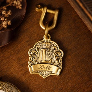 May include: A brass-toned metal pendant featuring a raised letter L, a small crown icon, and a banner with the engraved name Bella. The shield-shaped charm is attached to a U-shaped shackle connector with a smooth, matte finish.