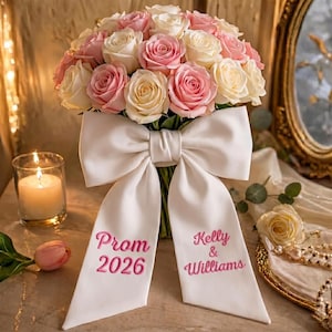 Könnte beinhalten: Ein Strauß aus rosa und weißen Rosen, gebunden mit einer großen weißen Schleife. Auf der Schleife sind die Worte "Prom 2026" und "Kelly & Williams" in Rosa gestickt. Eine brennende Kerze und eine rosa Tulpe sind im Vordergrund.