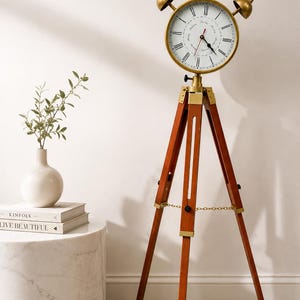 May include: A floor clock featuring a round white face with black Roman numerals and a brass-toned frame, mounted on a tall, three-legged wooden tripod stand with brass accents and a small chain connecting the legs for stability.