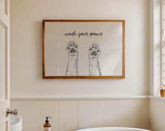 May include: A framed print with the text "wash your paws" above a sketch of two dog paws. The artwork has a brown wooden frame and is hanging on a cream-colored wall in a bathroom setting.
