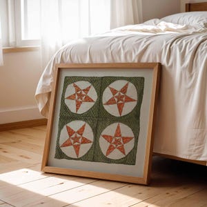 May include: A framed artwork with four star designs. Each star is red and white, on a green background within a white circle. The frame is light wood, leaning against a bed and wooden floor.